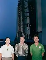 Three men stand in front of a rocket