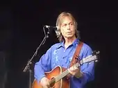 A man wearing a blue dress shirt, standing behind a microphone stand and holding a guitar.