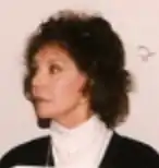 A white woman wearing a white turtleneck and a black jacket; her hair is dark and feathered in the style of the 1980s