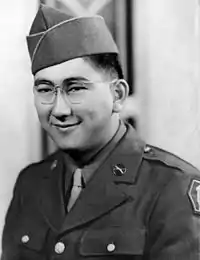 Head and shoulders of a smiling young man with dimples and round wire-framed glasses wearing a garrison cap and a military jacket over a shirt and tie