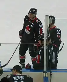 Hockey player in black uniform with a red leaf in the middle. He smiles and stands near the sideline of the rink.