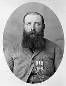 A black and white head and shoulders photograph of a balding white man with a full, dark, beard and a heavy wool coat
