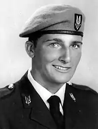 A studio photograph of a young, clean-shaven soldier, smartly turned out in his uniform. The flaming sword insignia of the Special Air Service is visible on each lapel of his jacket and prominently on his beret, above his left eye. He smiles into the camera with a look of assured confidence.