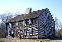 A modern photograph of an old, unadorned two story house clad in weathered shingles