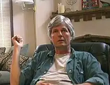 A casually dressed, middle-aged white man with wavy gray hair sits in an overstuffed chair in a living room