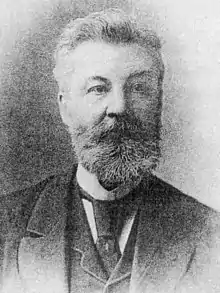 Head and shoulders of a bearded man wearing a three-piece suit, tie and high collar