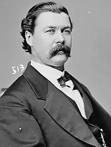 A man with black, wavy hair and a mustache wearing a black jacket and tie and white shirt