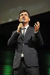 Oliver, wearing a black suit, talking into a microphone and gesturing with his hands.