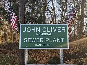 Photo of a green highway sign with "John Oliver Memorial Sewer Plant Danbury, CT" in large text. American flags hang off the corners of the sign. The sign is in a wooded area.