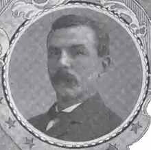 Portrait of a man with a drooping mustache and a dark suit coat, in a circular frame.