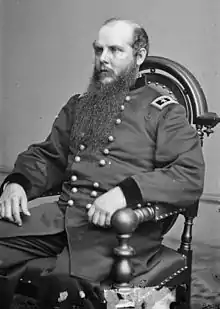 Photo shows a seated man in a dark military uniform with the two stars of a major general on his shoulder tab. He is balding and has a very long curly beard.