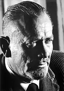 A photo of John Steinbeck. His hair is slicked-back and closely shaved on the sides. He has a mustache and facial hair on his chin.