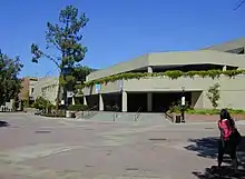 A photograph of a grey, three-story building on campus. The second story is offset back from the first and the third is offset form the second. The building has plants hanging over the second story onto the front of it. It is decorated with three unreadable banners. There are eight steps that lead to its covered entrance.