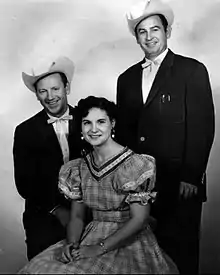 Two men wearing dark suits and cowboy hats standing either side of a woman wearing a gingham dress