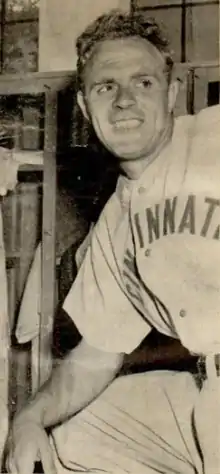 A man wearing a baseball uniform with "Cincinnati" written across the chest stands leaning on a propped up knee and smiles as he looks–the right.