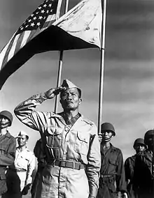 Filipino male in World War II Army Khaki uniform saluting in the foreground. A color guard in the background; the 48-star United States flag on the viewers left, a reversed/war Philippine Commonwealth flag on the viewers right.