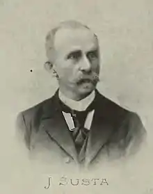 Josef Šusta, with a moustache and receding hairline and depicted in a black cravat and suiting. He is looking to the left of the camera. The image is of low quality, with no background being visible, and beneath it is the text "J Susta".