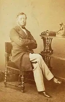 sepia photograph of a bearded man sat in a chair with his arms folded and legs crossed.