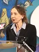 Color photo of an elderly women at a podium, speaking into a mic.