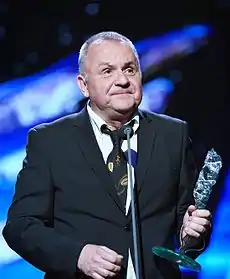 Jožo Ráž wearing a suit, standing in front of a microphone onstage, holding a trophy
