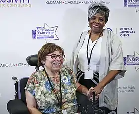 A photograph of Judy Heumann in her power chair next to Barbara Ransom. They are holding hands and smiling, standing in front of a sponsor banner.