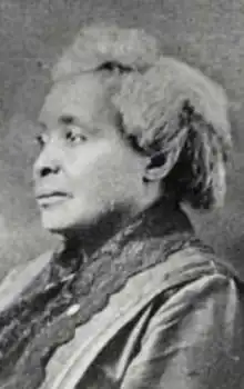 An older African-American woman with grey hair dressed back from her face, wearing a dress or jacket with dark lace trim