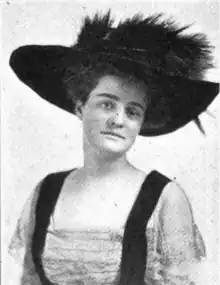 Photograph of a white woman wearing a large black hat, and a dress with an deep and wide square neckline