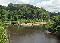 A body of water, seen from slightly above, that appears to fork in the center of the image. A metal bridge is partially visible crossing the right fork. The water in front is brownish, shading to green near the far shore. Some people are standing and fishing at the water's edge.