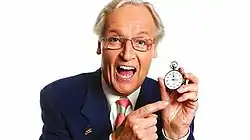 Image shows Nicholas Parsons smiling whilst holding a stopwatch in his left hand and pointing to the watch with his right hand