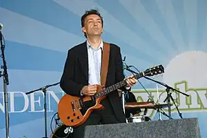 Justin Adams wearing a light patterned shirt, dark blazer, and dark pants, standing onstage, playing guitar, with drummer in background