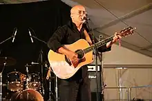 Bald Middle Eastern man wearing glasses and black shirt and pants, standing on stage, holding a guitar, and singing into a microphone in front of a drum kit.