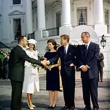 The men wear dark suits. A Naval officer in khaki stands behind them. The White House is in the background.