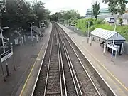 The view back from the footbridge. The pair of 3rd-rail-electrified tracks curve to the left between the 2 platforms. the left one has a sign with the number 2, stairs & a help-point. They both have a shelter, a ticket machine, display boards & lamp-posts. The trees on the left are darker in colour than the trees on the right. There is a silver car on the right & a modern office building.
