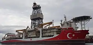 Medium sized ship viewed from starboard. Painted mostly red with the Turkish crescent and star in white near the bow. Above the bow is a helicopter deck and there is a large derrick amidships. Two small cranes are built into the side of the ship.