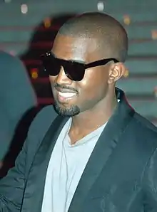 An Afro-American man with a ponytail on top of her head is wearing a black leather jacket before a light-coloured background.