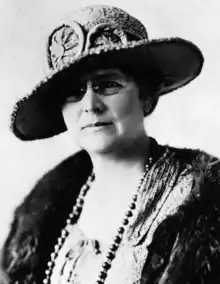 A middle-aged white woman wearing an ornate hat with a broad brim, glasses, a fur stole, and beads.