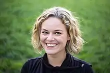 Katherine Maher in 2016. She is seen with light skin, blonde hair, and blue eyes. She is seen wearing a black shirt.