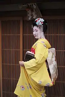 A young apprentice geisha stood outside a traditional Japanese building. Her yellow kimono's sleeves are shorter than a maiko's, and she wears more hair accessories than a maiko would.