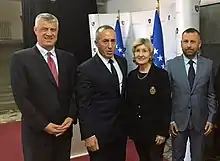 Three men and one woman in suits stand on a red carpet in front of two blue flags.