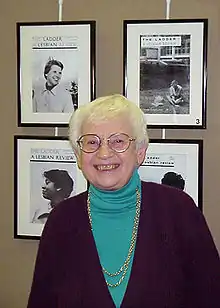 Color photo of smiling older white woman with short white hair, wearing a green turtleneck and brown cardigan with gold chains; she is standing in front of four framed black-and-white images against a taupe wall
