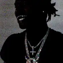 A low quality close-up shot of Ken Carson smiling as he stands in front of a gray backdrop; Carson is wearing a black t-shirt, diamond chains, and an earring.