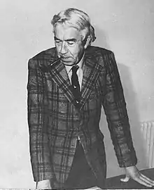 Kenneth Williamson is standing, leaning on a desk. He is wearing a suit and has a pipe in his mouth.