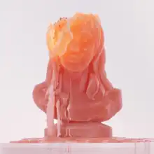 A red-orange candle representing Kesha's head is positioned on top of a surface. The candle wax is melted, disfiguring the top of the "head".