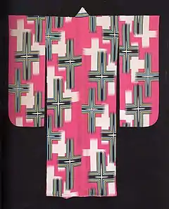 A sugar-pink, long-sleeved kimono viewed from the back. The kimono is lined in a lighter shade of pink. It is decorated with a stencil-printed design of elongated white and blue crosses, the four ends of each cross tapering off in a manner reminiscent of a rough brush stroke.