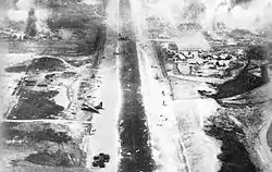 Black and white image from the rear of a plane showing a landing strip littered with damaged aircraft