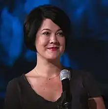 Woman with short, dark bobbed hair, a dark shirt, and a small necklace stands in front of a microphone against a blue backdrop.