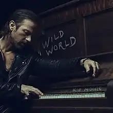 A man wearing a black leather jacket, sitting next to a piano. The album title is next to him and the artist's name appears above the keys, both written in chalk-like drawing.