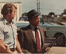 Kirk Douglas (right) holding a gun and John Schneider (left) looking at Douglas, with two light blue trucks in the background, both seen on the set of the movie Eddie Macon's Run during its filming in 1983