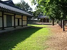 On a sunny day, a Korean traditional wooden building painted with white and dark red stands on a grass field. Luxuriant trees are seen on the right while a gate is shown at a distance.