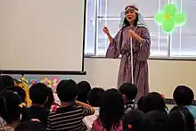 A photograph of a woman wearing a red-and-blue-striped robe holding a white stick and speaking while standing in front of a group of children who are facing her